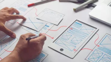 Person zeichnet Wireframe-Designs auf Papier mit einem Tablet daneben.